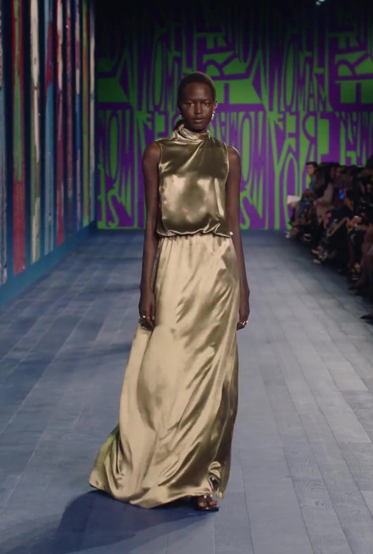 Modelo da Dior usando Vestido longo de cetim dourado, sem mangas, usado por modelo na passarela com fundo de grafite. Elegante e chamativo.