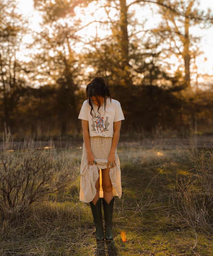 Menina usando camiseta estampada country, saia longa florida e botas de cano médio na natureza.
