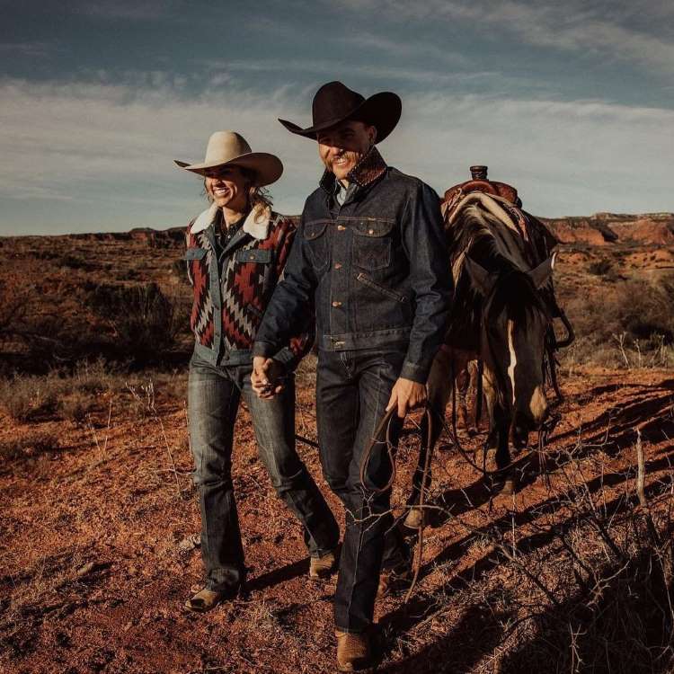 Casal andando em local com terra, levando seu cavalo. Eles usam roupas no estilo cowboy, moda country, com peças em jeans e chapéu.