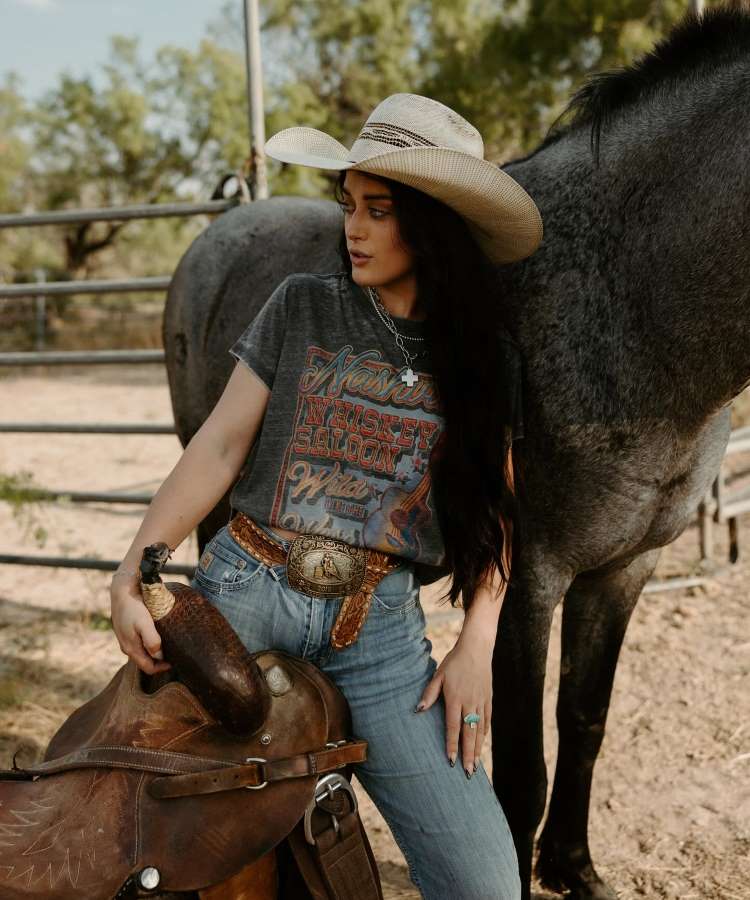 Mulher segurando cela ao lado de um cavalo, usando camiseta estampada country, calça jeans, cinto com fivela e chapéu.