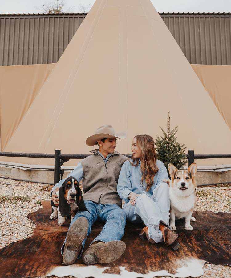 Casal com seus dois cachorros sentados em um tapete de couro no chão de pedra. Os dois usam roupas jeans e sapatos country.