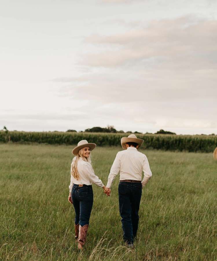 Casal de costas andando no mato, usando camisa branca, chapéu, calça jeans, cinto e bota western.