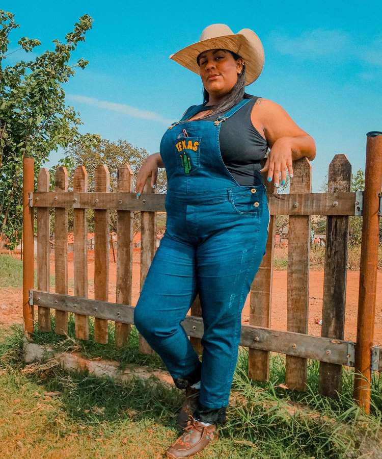 Mulher apoiada em cerca de madeira, usando regata preta, macacão jeans com estampa do Texas, sapato de couro e chapéu de cowboy.