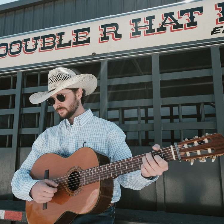 homem usando camisa xadrez branca, calça jeans, cinto com fivela e chapéu de cowboy, segurando violão