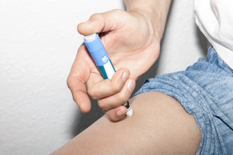 Pessoa de pele clara usando shorts azul, aplicando medicamento em caneta na perna