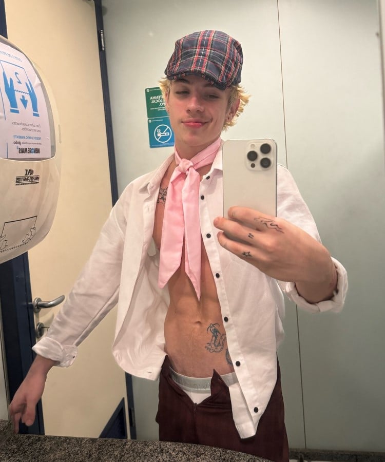 Juliano Floss em selfie, usando camisa aberta, lenço rosa no pescoço, boina e calça com zíper aberto, ele está em um banheiro público
