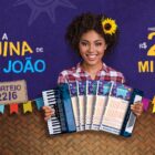 Imagem de fundo azul com mulher de pele negra usando camisa xadrez branca e vermelha, com uma sanfona de bilhetes da Quina de São João, além de informações sobre a loteria, como data (22 de junho) e prêmio (R$ 220 milhões)