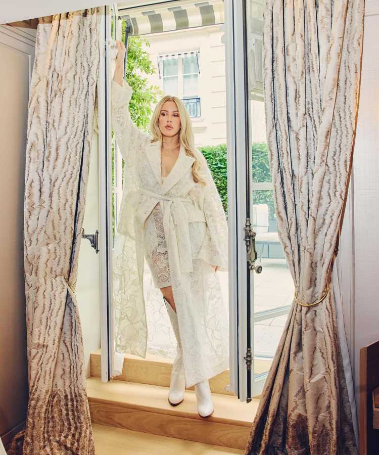  Ellie Goulding usando Conjunto branco com robe de renda, calça e sapatos brancos. Look elegante e sofisticado, ideal para ocasiões formais.