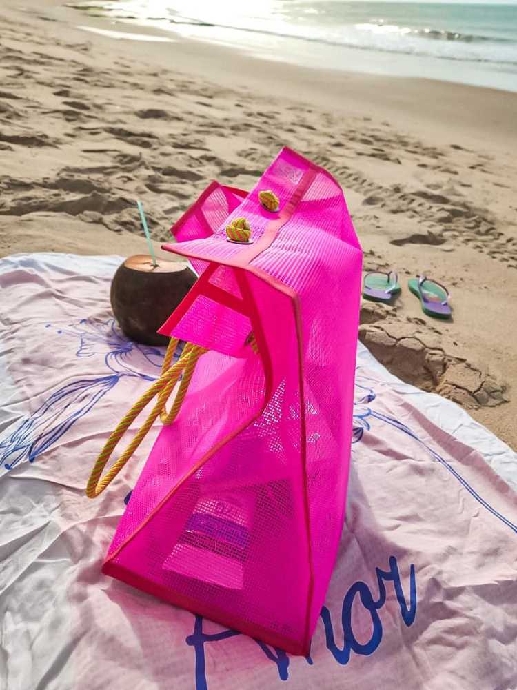 Foto de bolsa de praia rosa com alça amarela, sobre canga branca, ao lado de coco e na areia