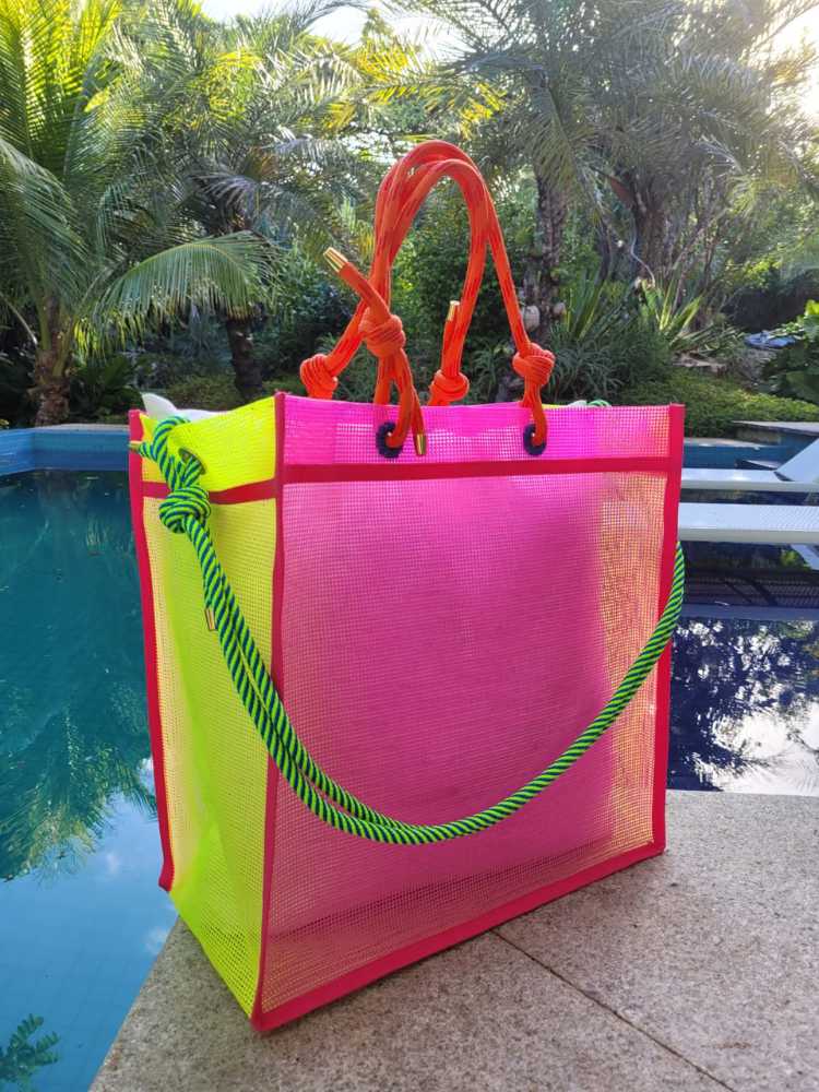 Bolsa de praia/piscina nas cores rosa e amarela, com alças em verde e vermelho. A bolsa está na beirada de uma piscina, com árvores atrás