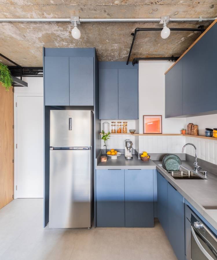 A imagem mostra uma cozinha moderna com um interior minimalista. O esquema de cores consiste em tons suaves, com armários azul-acinzentados e um teto de concreto. Há uma geladeira de aço inoxidável à esquerda, ao lado de armários altos da mesma cor. Os armários inferiores estão sob uma bancada branca que abriga uma pia com torneira e alguns itens de cozinha, como pratos e potes. Na bancada, também há dois limões, uma laranja e uma garrafa de vidro com líquido. Acima da bancada, há duas prateleiras; uma com copos e garrafas e outra com plantas verdes. O piso é claro, possivelmente de azulejo ou laminado. Quatro luminárias pendentes iluminam o espaço junto com a luz natural que entra de fora da imagem.