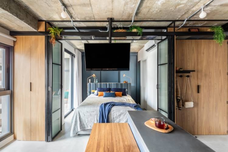 A imagem mostra um quarto moderno com uma estética de design industrial. O quarto possui um teto de concreto exposto com canos e dutos visíveis, pintados de preto. Uma grande cama está posicionada centralmente no espaço, com roupas de cama cinza e almofadas azuis e laranjas que adicionam um toque de cor. Acima da cama, há uma grande TV de tela plana contra uma parede escura, criando um ponto focal. À esquerda da cama, há portas de madeira de altura total, provavelmente armários, que complementam o piso de madeira que se estende por todo o espaço.À direita do quarto, há uma longa mesa ou bancada de madeira que corre paralela à cama, sobre a qual repousam itens decorativos, incluindo o que parece ser uma bandeja com copo. O quarto é bem iluminado pela luz natural que entra por janelas não visíveis na imagem, mas sugeridas pelos reflexos nas superfícies e objetos dentro do quarto.