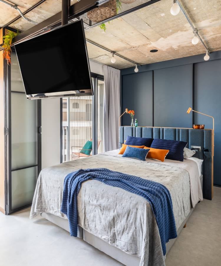 A imagem mostra um quarto moderno. A cama está centralizada, com um edredom cinza e várias almofadas em tons de azul e laranja. Acima da cama, há uma grande TV de tela plana montada no teto, inclinada para baixo em direção à cama para facilitar a visualização. À direita da cama, há uma luminária de chão alta com acabamento dourado, adicionando um toque de elegância ao ambiente. O quarto tem teto de concreto com dutos expostos, conferindo um estilo industrial, enquanto as paredes são pintadas em um tom calmante de azul.À esquerda da cama, há uma porta de vidro que parece levar a outro cômodo ou talvez a uma varanda, permitindo a entrada de luz natural. De um lado dessa porta de vidro, há plantas em vasos penduradas no teto, introduzindo um toque de verde ao espaço.