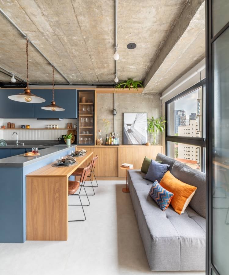 A imagem mostra um interior de apartamento moderno com um layout de plano aberto. O teto é de concreto inacabado, e há duas luminárias pendentes com design simples. À esquerda, há uma área de cozinha com armários azul-escuros e detalhes em madeira. A bancada da cozinha também é de madeira, e há vários bancos com assentos pretos e pernas de madeira ao longo dela. Acima da bancada, prateleiras abertas seguram vários itens de cozinha.Ao lado da cozinha, há uma área de jantar com uma mesa de madeira e cadeiras com estofado laranja e estruturas pretas. Uma grande janela ocupa quase toda a parede direita, proporcionando muita luz natural e uma vista dos prédios do lado de fora.Em primeiro plano, há uma área de estar com um sofá cinza adornado com almofadas de padrões geométricos em tons de azul e laranja. Em frente ao sofá, há um tapete em cores suaves e, à direita, uma pequena mesa lateral com itens decorativos.