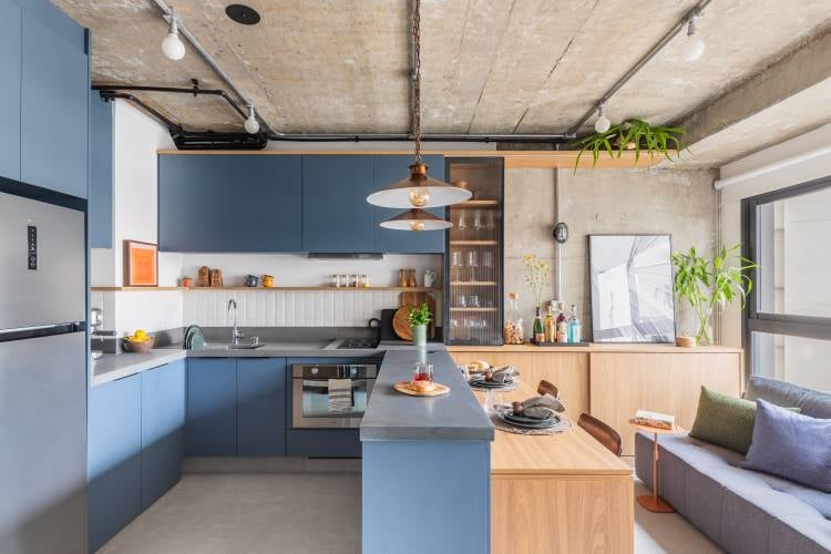 A imagem mostra uma cozinha moderna e uma área de estar em um layout de plano aberto. A cozinha possui armários elegantes de cor azul escuro com acabamento fosco, complementados por uma bancada cinza clara. Uma ilha central com o mesmo esquema de cores serve como área de preparação e bar, equipada com dois bancos altos. Acima da ilha, há uma luminária pendente com um tom metálico quente.