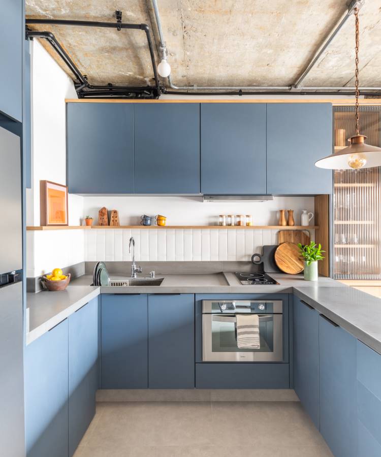 A imagem mostra uma cozinha moderna com um design minimalista. Os armários são de cor azul fosco, e as bancadas são cinza claro, criando uma estética de tons frios. A cozinha possui uma pia embutida com uma torneira alta e está equipada com eletrodomésticos de aço inoxidável, incluindo um forno. Acima da bancada, há um backsplash de azulejos brancos que adiciona textura ao espaço. À esquerda, há uma estante aberta exibindo vários itens de cozinha e obras de arte emolduradas. Uma luminária pendente com acabamento em madeira natural está suspensa acima, contribuindo para o ambiente contemporâneo.