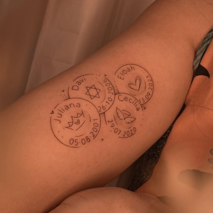 Braço de pessoa de pele clara tatuado com medalhas com nomes, símbolos e data de nascimento 