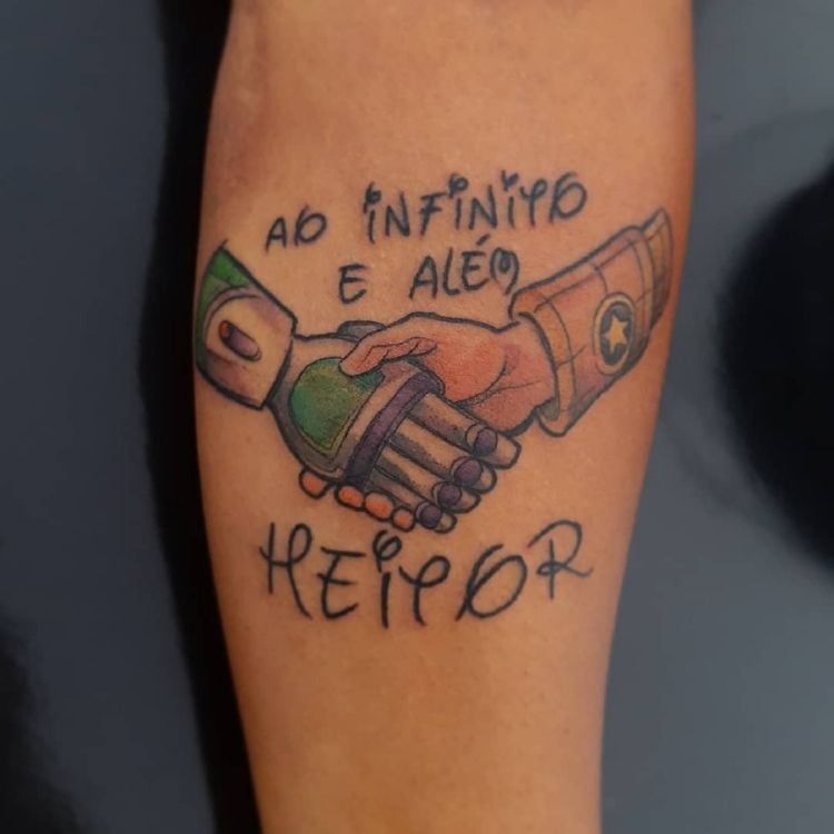 Tatuagem de desenho de mãos do Woody e Buzzlightyear com frase "ao infinito e além" e nome "heitor"