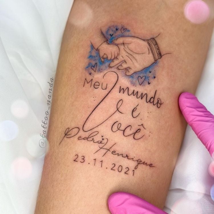 Foto de tatuagem de mão grande e pequena dadas, frase "Meu mundo é você, Pedro Henrique" e data de nascimento
