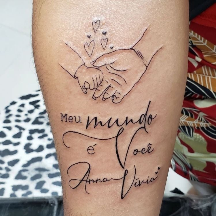 Foto de tatuagem de mão grande sendo segurada por mão de bebê, frase "Meu mundo é você" e nome "Anna Vívia"