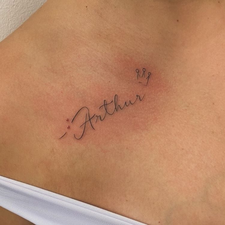 Foto de tatuagem escrito "Arthur" com uma coroa em ombro de pessoa de pele clara 