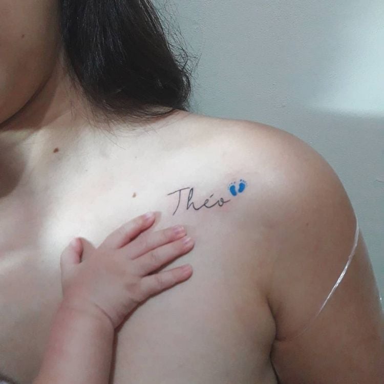 Mulher de pele clara com mão de bebê no colo e tatuagem escrito "Theo" com pés pequenos azuis