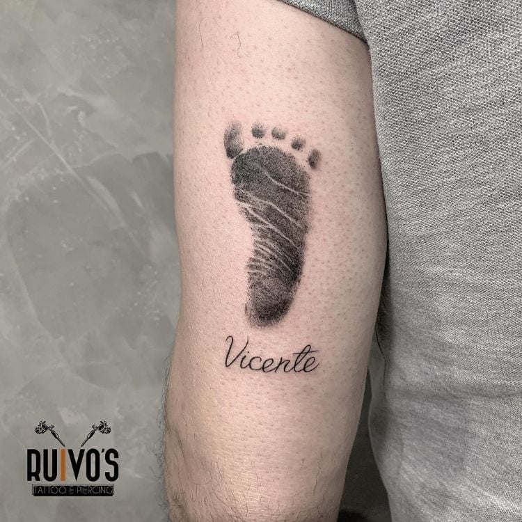 Braço de homem de pele clara tatuado com impressão de pé e nome "Vicente"