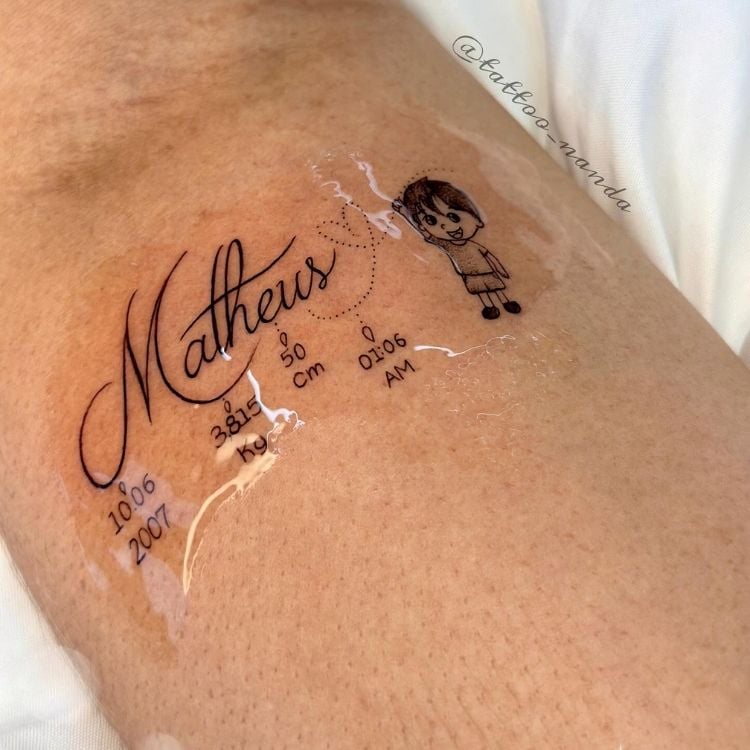 Tatuagem em braço de pessoa de pele clara com nome "Matheus", varal com informações de nascimento e desenho de menino