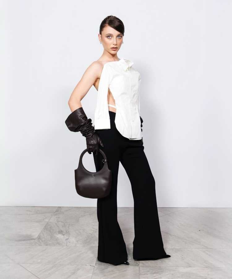 Emma Chamberlain usando Calça preta flare, camisa branca desconstruída, uma luva preta longa e bolsa preta. Look moderno e elegante.