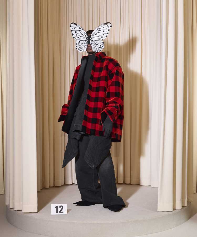 Modelo da Balenciaga usando Camisa xadrez vermelha e preta, jeans escuros, blazer preto e máscara branca de borboleta. Look ousado e estiloso na Semana de Alta Costura de Paris inverno 2024