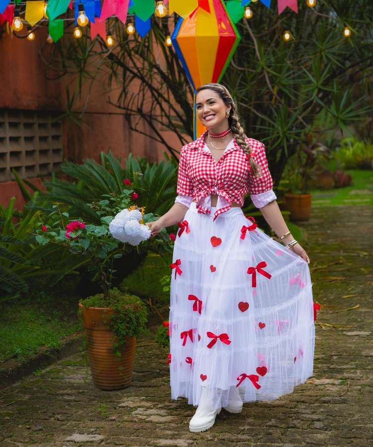 mulher com look junino, saia branca com laços e corações vermelhos. Ela usa uma camisa vermelha xadrez