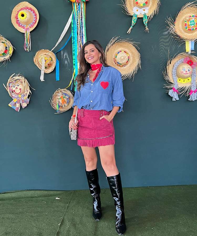 Mulher com look junino composto com camisa xadrez, saia, botas de cano longo e laço no pescoço