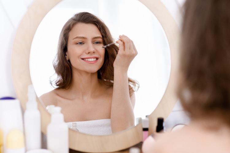 mulher sentada em sua penteadeira olhando para o espelho enquanto aplica retinol no rosto