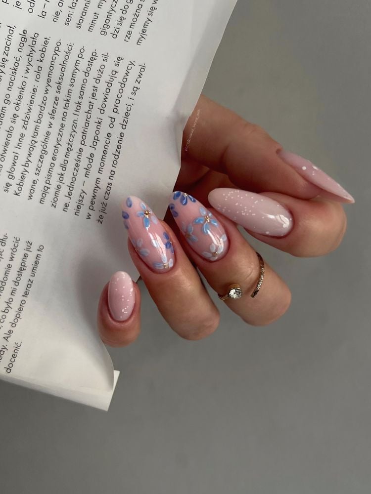 Foto de mão de pessoa de pele clara segurando jornal, com unhas almond em rosa clarinho e nail art de flores azuis no anelas e médio