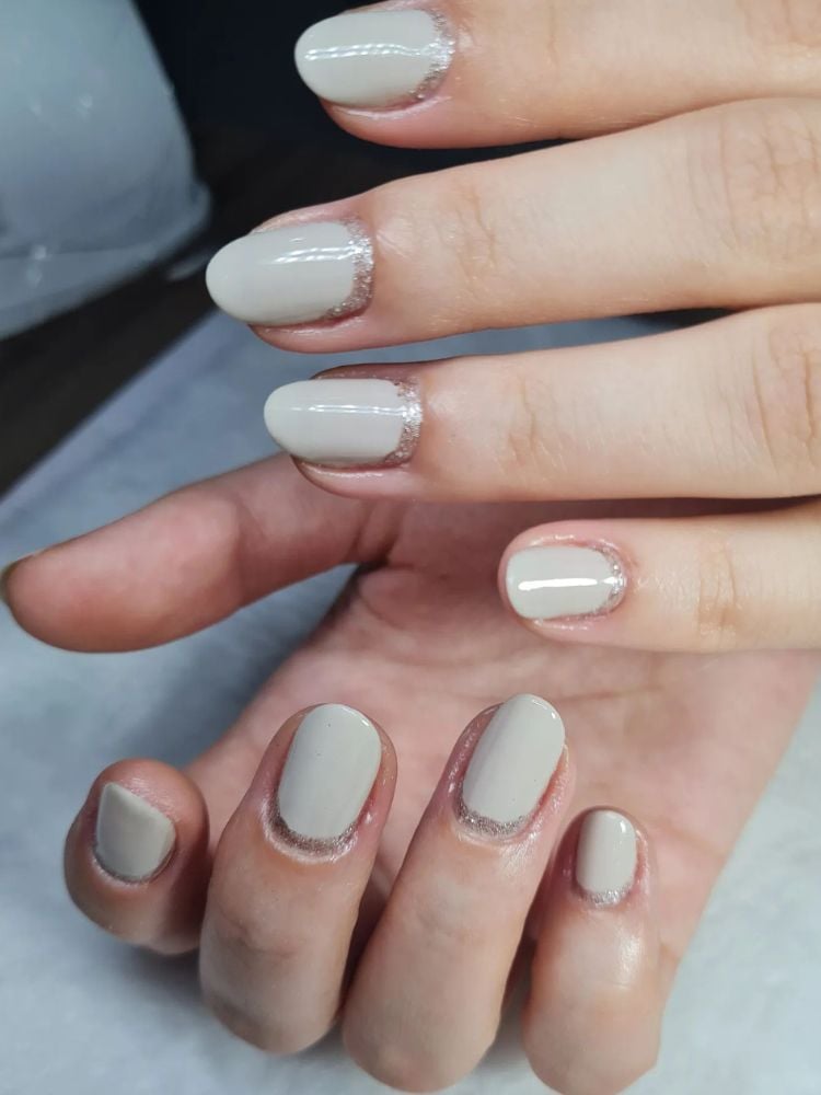 Foto de mãos de pessoa de pele clara usando esmalte cinza claro e francesinha invertida com glitter
