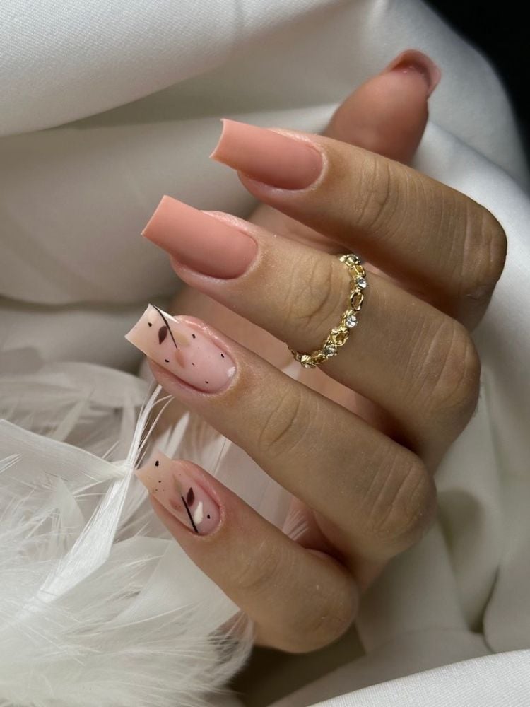 Foto de mão de pessoa de pele clara com unhas quadradas em tom de nude, sendo o anelar e dedinho decorados com desenho de ramos 