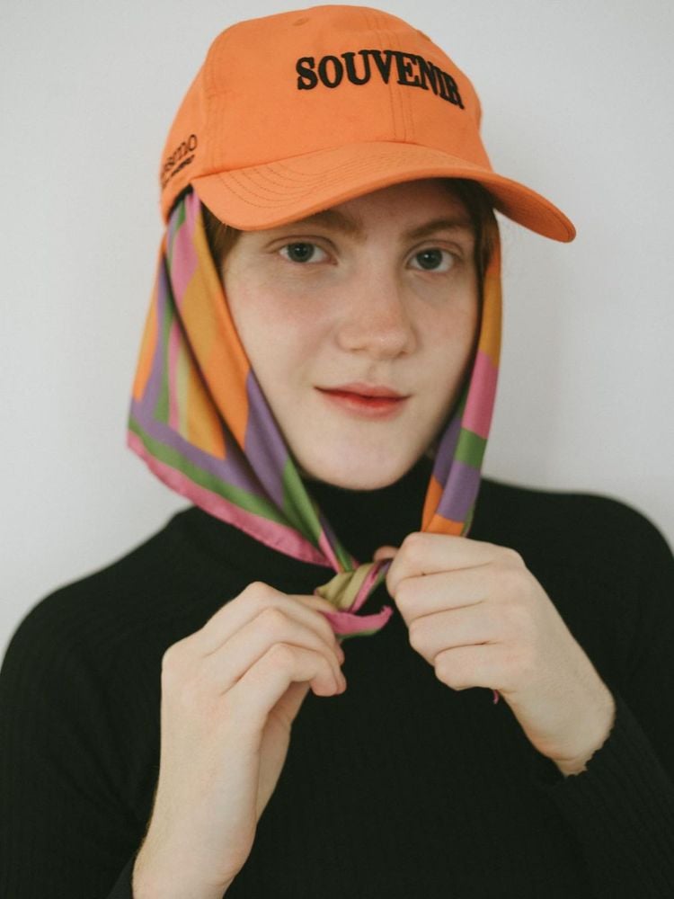 Foto de mulher usando boné laranja com lenço colorido por baixo