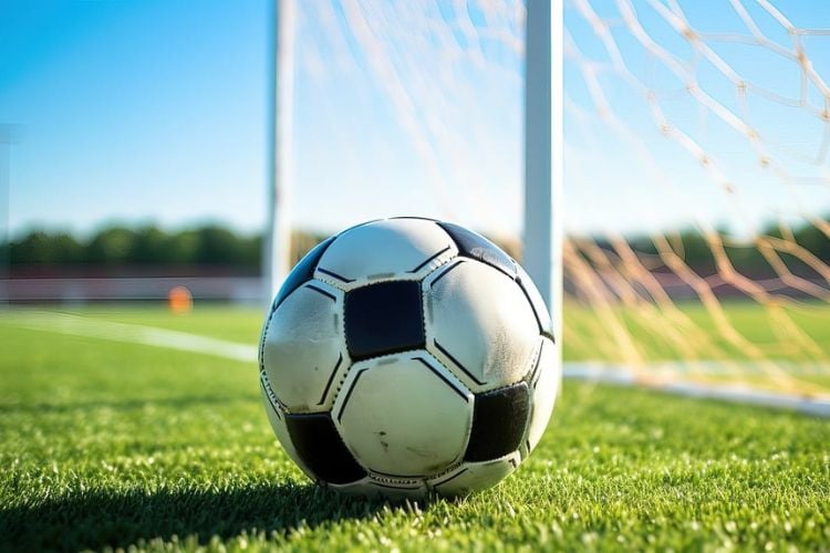 Foto de bola de futebol branca e preta em campo de grama sintética, ao lado de rede do gol