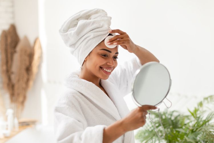 Mulher de pele negra usando toalha branca e roupão branco enquanto segura um espelho e limpa o rosto