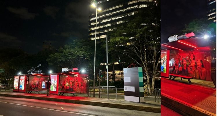 A imagem mostra uma cena urbana noturna. À esquerda, há um ponto de ônibus bem iluminado com estruturas vermelhas e anúncios com pessoas. Ao fundo do ponto de ônibus, vemos prédios altos com janelas iluminadas. Uma árvore sem folhas também é visível atrás do ponto de ônibus. No centro da imagem, há um poste de luz e o que parece ser um painel de informações ou um poste indicativo. À direita, temos outra vista do mesmo ponto de ônibus, focando no telhado vermelho e no painel lateral com mais anúncios.