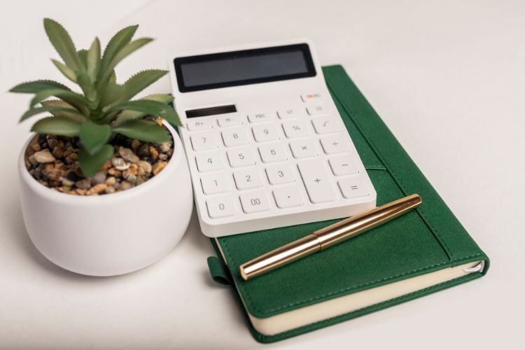 Tampo de mesa branco com caderno de couro verde, caneta dourada, calculadora e vaso de planta branco com suculenta e pedras