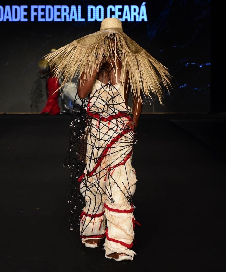 Um modelo desfila no evento, vestindo um traje com estrutura de rede sobreposta a tecidos brancos e vermelhos. O destaque é um grande chapéu de palha que cobre a cabeça e os ombros do modelo. 