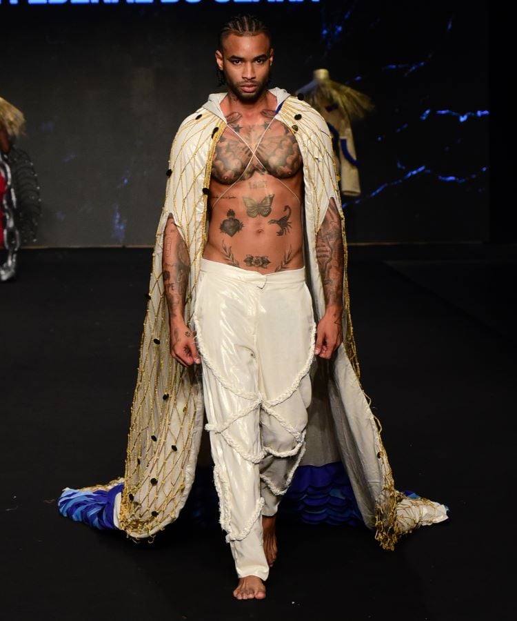 Um modelo masculino desfila na passarela durante o evento “UFC no Concurso dos Novos” no DFB Festival 2024. Ele veste uma roupa branca com detalhes dourados e azuis, incluindo uma capa longa e calças largas. O estilo da roupa é contemporâneo e étnico.