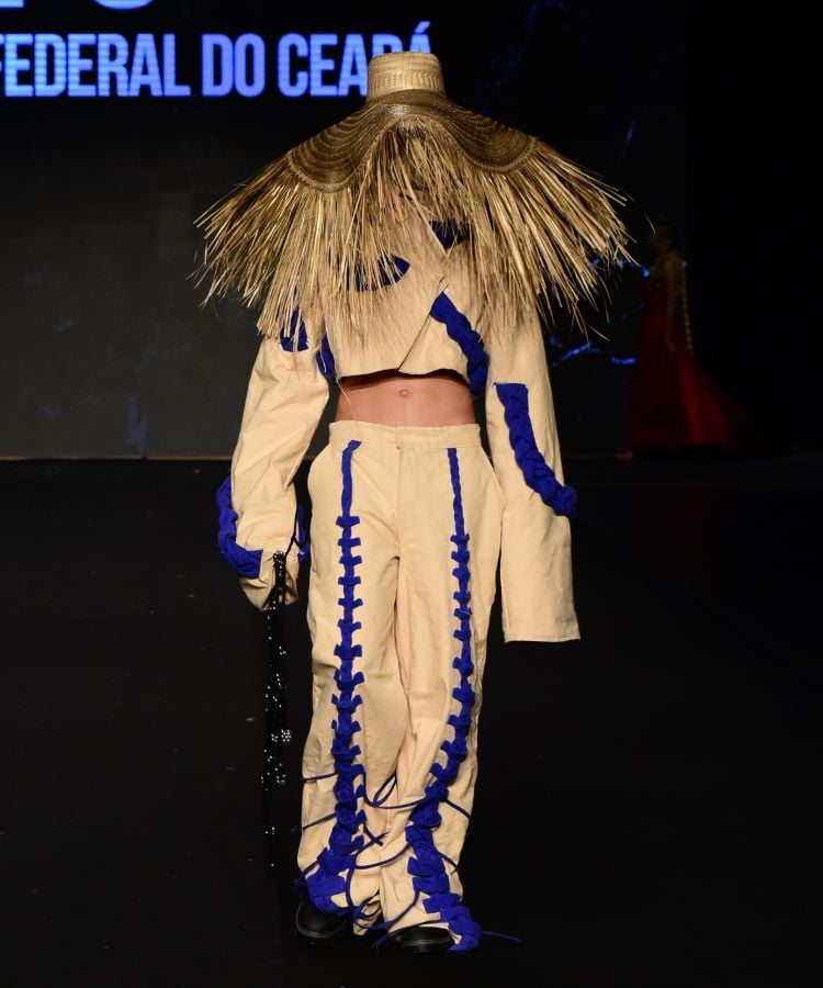 A foto mostra um modelo vestindo uma roupa bege com detalhes azuis e um chapéu de palha grande que cobre toda a cabeça. O evento é parte do DFB Festival 2024 e envolve estudantes da UFC (Universidade Federal do Ceará).