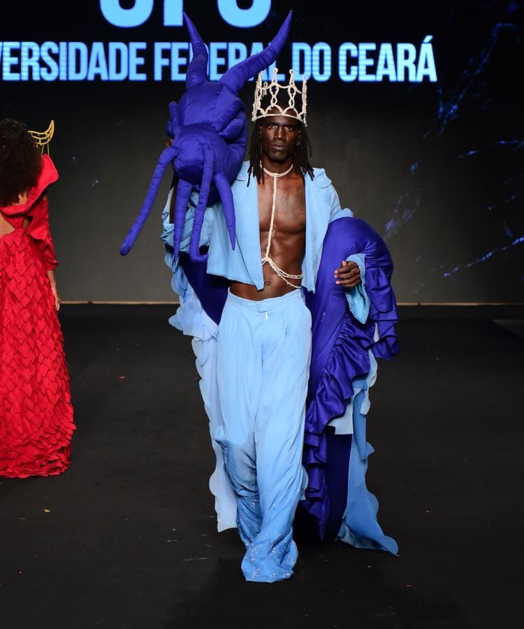 Um modelo masculino desfila na passarela usando uma roupa azul vibrante com detalhes em roxo. Ele usa luvas longas, uma capa e uma coroa, com o torso exposto. As luzes destacam sua postura confiante.