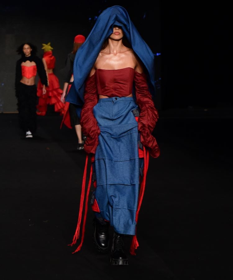  Uma modelo desfila no DFB Festival 2024 usando uma peça criada pelo SENAI Moda RN. A roupa combina texturas e cores, com destaque para o azul e vermelho. A modelo usa uma capa azul sobre a cabeça, um top vermelho e uma saia longa azul com detalhes em vermelho. Outros modelos aparecem desfocados ao fundo. 