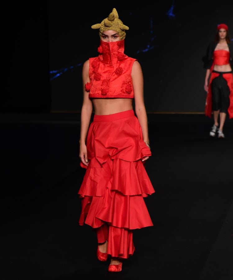 Uma modelo desfila com elegância em um conjunto vermelho vibrante, composto por um top e uma saia de babados em camadas. O evento acontece no SENAI Moda RN durante o DFB Festival 2024. A passarela tem um fundo escuro, destacando a moda contemporânea.