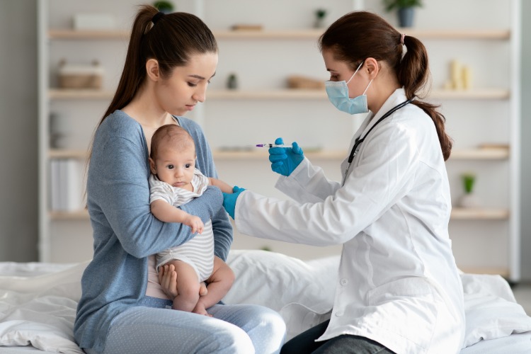 Enfermeira de pele clara usando jaleco e luvas aplicando vacina em bebê de pele clara, usando body branco, no colo da mãe de calça e cardigan azuis