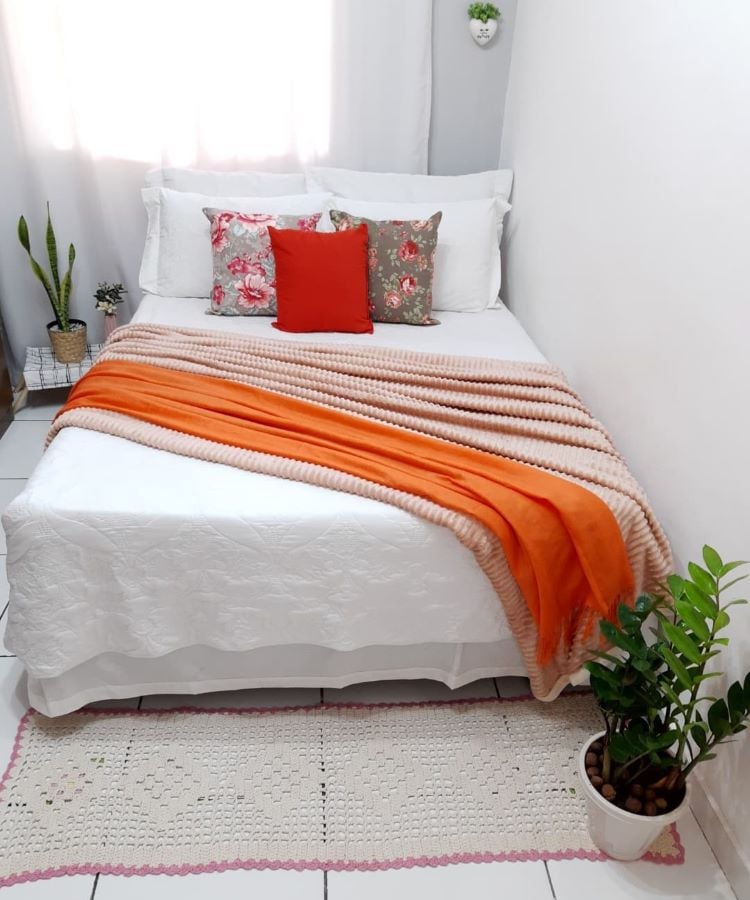 A imagem mostra uma cama bem arrumada em um quarto iluminado. A cama tem roupas de cama brancas e está decorada com vários travesseiros: dois com padrões florais e um sólido vermelho no centro. Um cobertor laranja com uma textura padronizada está colocado aos pés da cama, adicionando um toque de cor à roupa de cama branca. À esquerda da cama, há uma pequena planta verde em um vaso no chão, e outra planta maior está situada à direita da cama, também no chão. A parede atrás da cama tem uma decoração minimalista com apenas um pequeno item redondo visível, que pode ser interpretado como uma obra de arte ou uma luminária. Abaixo, há um tapete off-white com bordas rosa e detalhes de franjas parcialmente visíveis na borda inferior da imagem. 