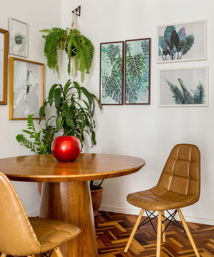 A imagem mostra uma cena interior focada em uma pequena área de jantar. No centro, há uma mesa redonda de madeira com uma superfície lisa e polida. Sobre a mesa, há um vaso ou fruteira brilhante e vermelha. Duas cadeiras com estofado de couro marrom e pernas de madeira estão posicionadas ao redor da mesa, uma empurrada para dentro e outra ligeiramente puxada para fora, como se alguém tivesse acabado de se levantar.O piso apresenta um padrão intrincado de espinha de peixe feito de madeira clara e escura. A parede atrás do conjunto de jantar é adornada com obras de arte emolduradas; há seis quadros no total, exibindo várias ilustrações botânicas que incluem folhas verdes e motivos de plantas. Acima das obras de arte, há uma planta pendurada com folhagem verde exuberante caindo. No canto dessa parede, no chão, há outras plantas em vasos, dentre elas, uma sendo a zamioculca